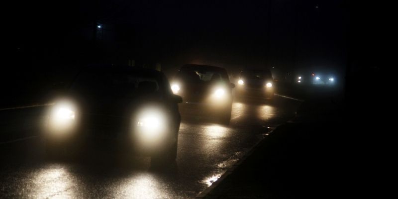Cars navigate a slick, illuminated road at night, reflecting the glow of streetlights on the wet surface.