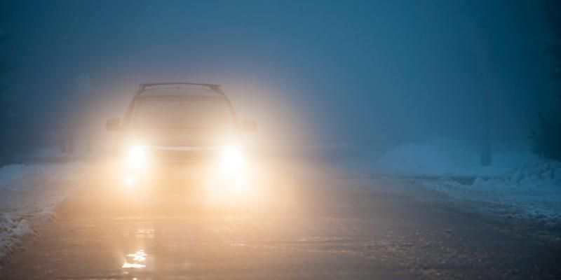 A car navigates a snowy road at night, illuminated by headlights against a backdrop of falling snowflakes.