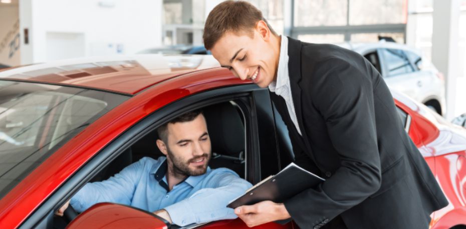Car salesperson showing a clipboard to a man sitting in a red car inside a dealership.