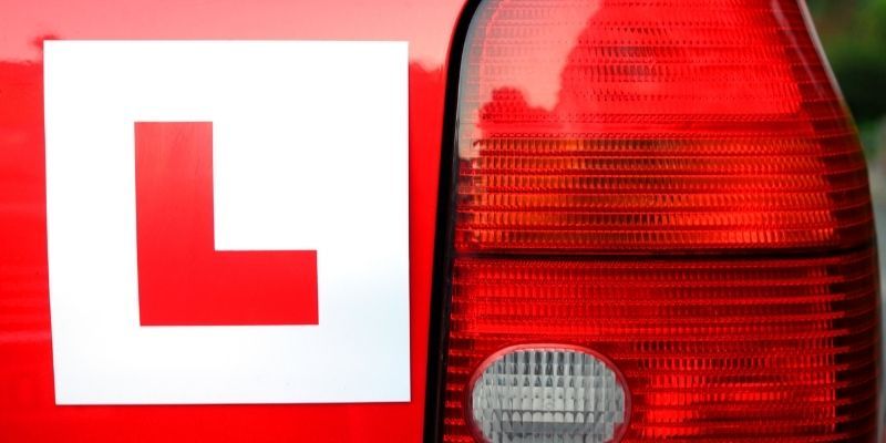 Red car with an 'L' learner driver sign on the back, indicating it is being used for driver training or by a new driver.