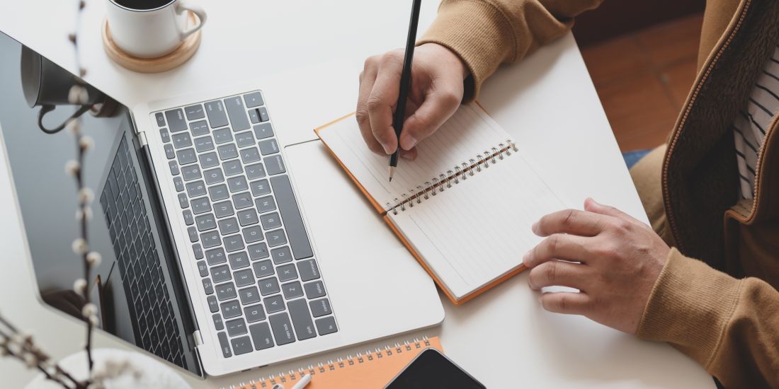 A hand poised over a notepad on a table with a laptop and coffee.