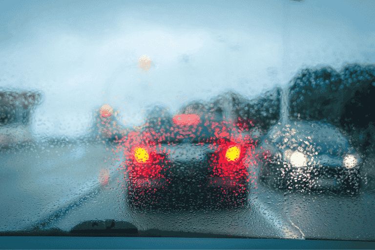 Rain-covered windscreen with blurred traffic ahead, highlighting glowing red brake lights through heavy rain and low visibility.