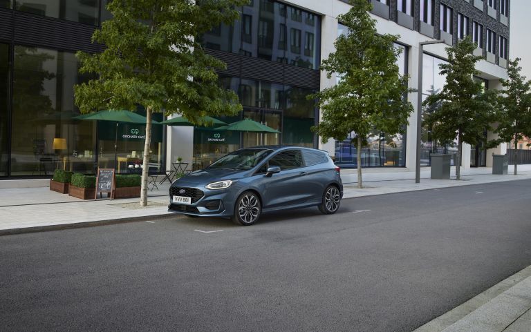 Grey Ford Fiesta hatchback parked on a modern city street outside cafés and apartments