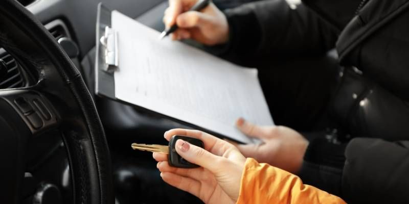 Someone holding a car keys in the drivers seat while another individual is writing notes on a clipboard