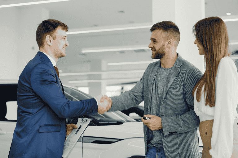 Young couple buying a car from a used car dealership, the salesman and gentleman buying the car are shaking hands.