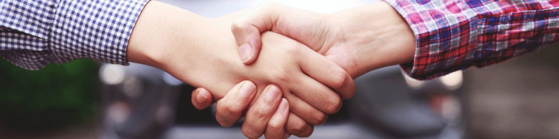 Close-up of two people shaking hands, symbolising agreement or car purchase, with blurred car in background.