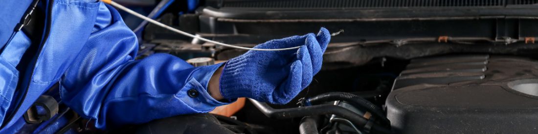 A person in a blue suit and blue gloves checking a cars dipstick in front of an open bonnet.