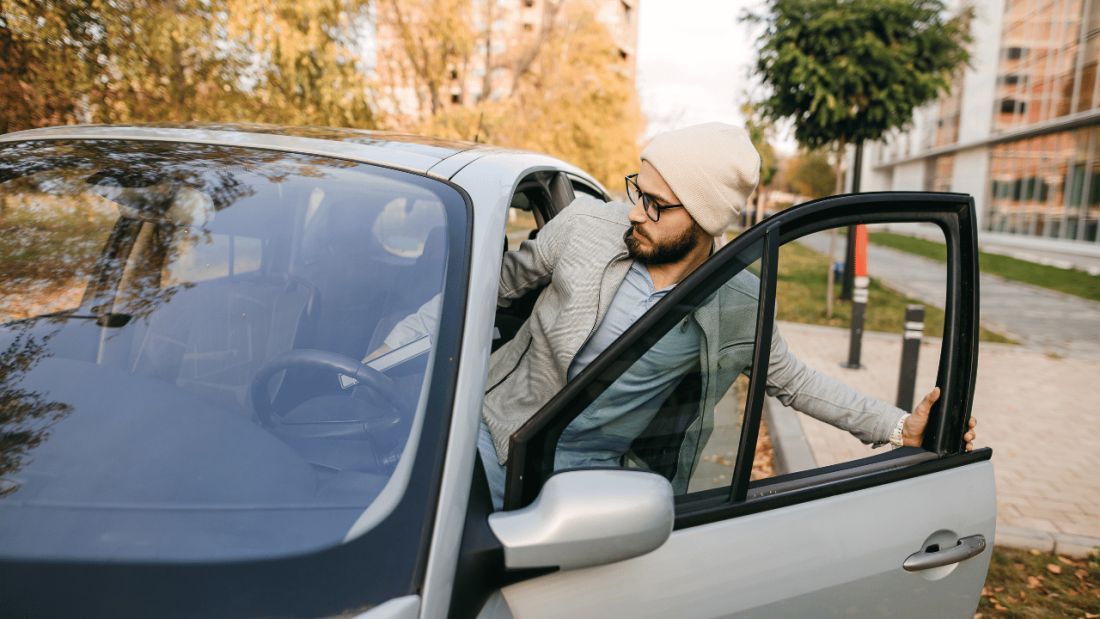A man in a grey jacket and a beanie hat with glasses on getting into a silver car.
