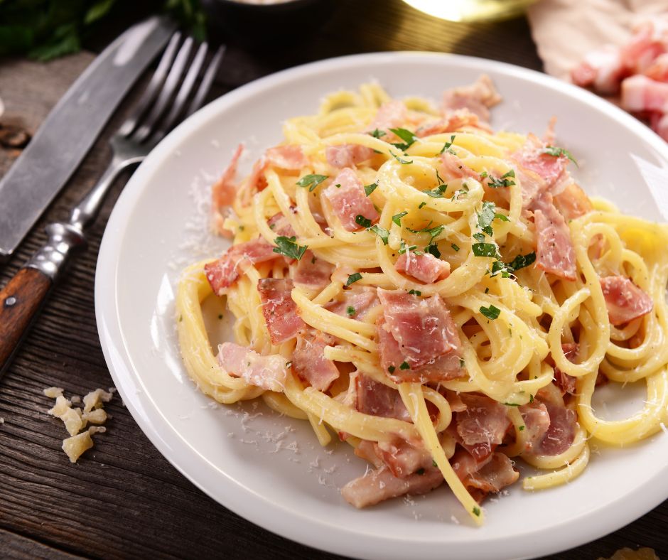 A plate of spaghetti carbonara with bacon bits. Cutlery has been placed on the left-hand side. 