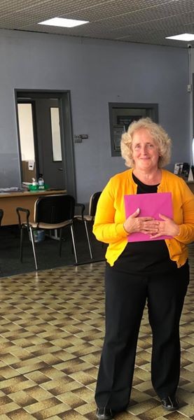 Annette is smiling holding a pink envelope to chest. She is wearing a black t-shirt and trousers and a yellow cardigan.