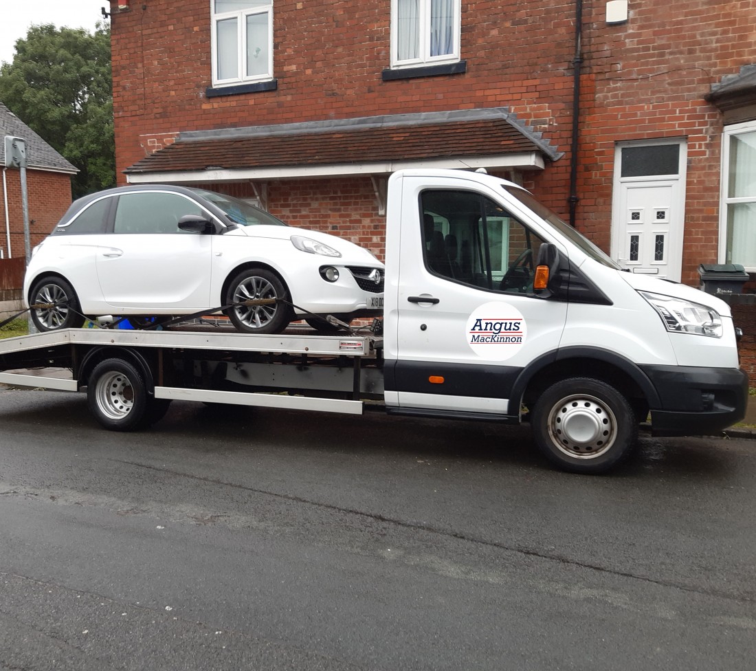 The white Angus MacKinnon transport van parked in front of a house with a small white car on the back.