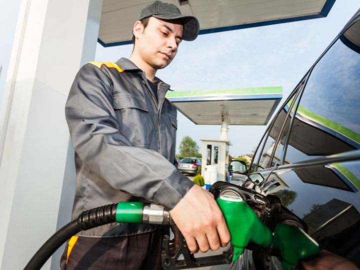 A man refuels his car at a petrol station, holding the fuel nozzle while the vehicle is parked nearby.