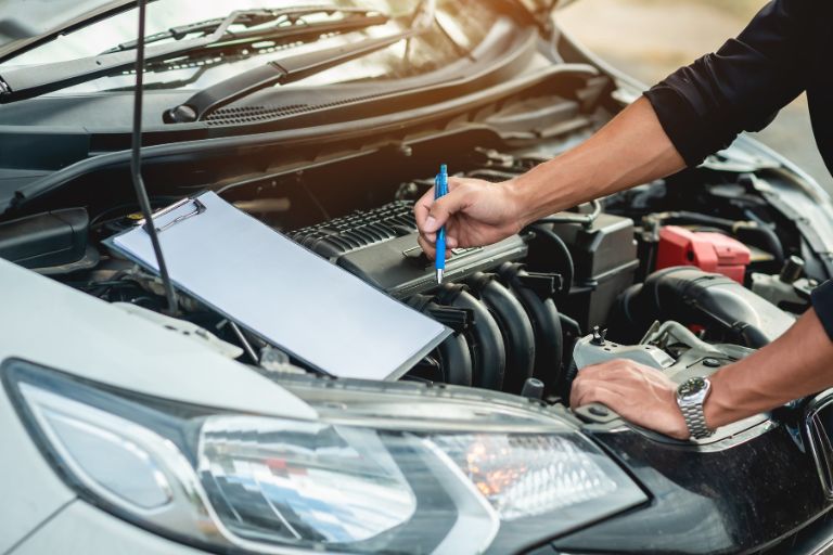 An exposed car engine and other parts. A clipboard is resting on the exposed mechanics and two hands are points to the vehicle elements to make an assessment.