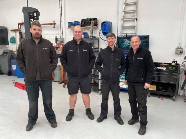 The Motor City Plymouth car servicing team, consisting of four men, standing together in front of their workshop.			 			