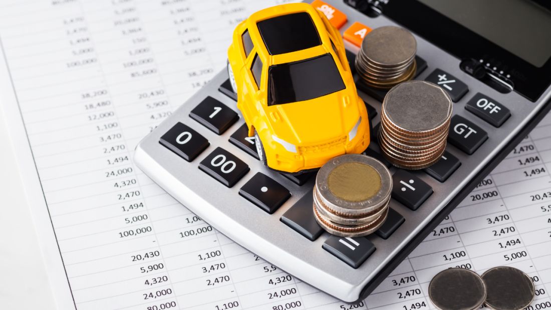 Yellow toy car placed on a calculator surrounded by stacked coins, representing car finance costs, budgeting, and vehicle affordability.			 			