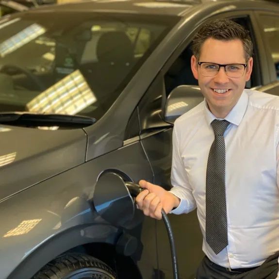 A gentleman with glasses and smart attire pluggin in an electrical charger into a grey car