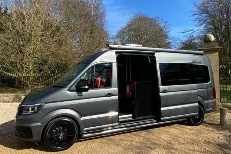 Silver camper van with sliding side door open, black alloy wheels, and red interior seats, parked outdoors on a sunny day.