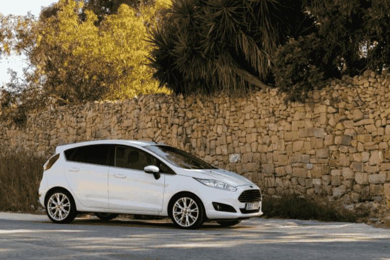 White Ford Fiesta parked beside a stone wall on a quiet road.