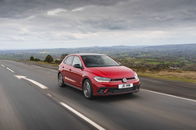 Red Volkswagen Polo driving on a road, under a stormy grey sky.