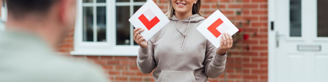 Learner driver holding L plates during driving lesson outside home.